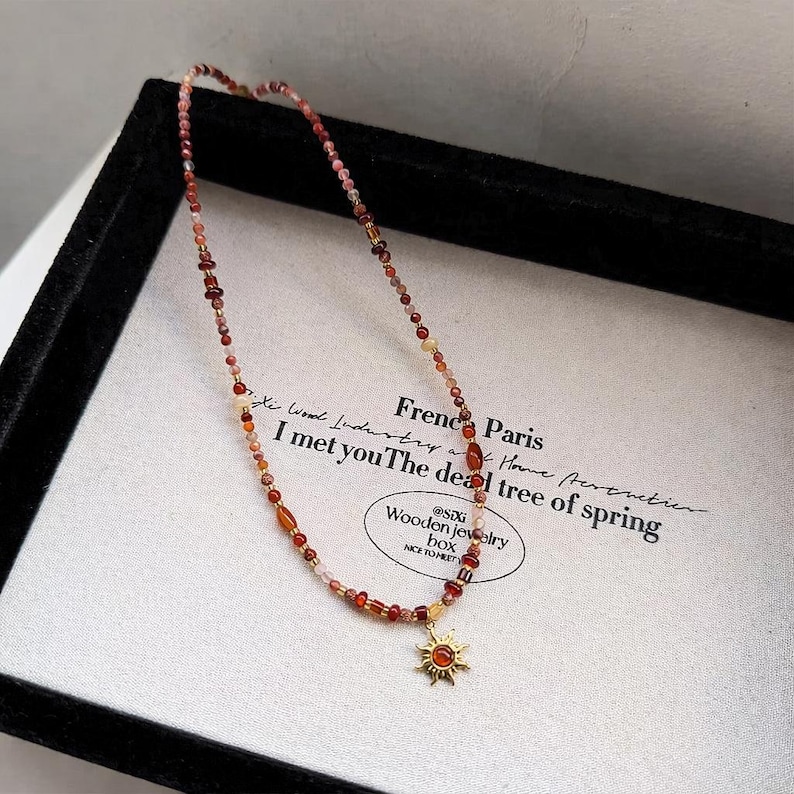 May include: A beaded necklace with a sun pendant. The necklace features small, colorful beads in shades of red, orange, and pink. The sun pendant is gold-colored with a red center. The necklace is displayed on a white surface with text.