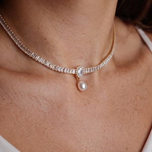 May include: A gold-toned necklace featuring a continuous row of rectangular baguette-cut clear stones. The center of the necklace displays a small teardrop-shaped pendant with a single white pearl suspended from a halo of tiny clear stones.