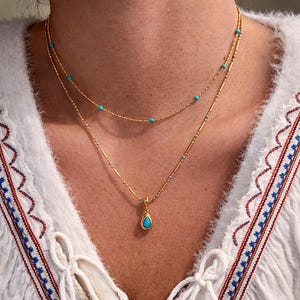Puede incluir: Collar dorado de varias capas con detalles turquesa. La capa superior presenta pequeñas cuentas turquesas, mientras que la capa inferior tiene un colgante en forma de lágrima con una piedra turquesa. Se lleva sobre una blusa blanca.