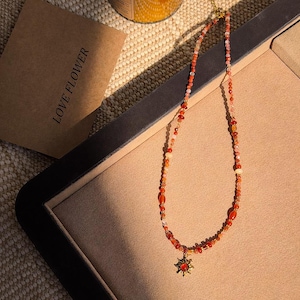 May include: A beaded necklace with a gold pendant in the shape of a sunburst. The necklace features a variety of orange, red, and cream-colored beads. The necklace is displayed on a beige surface, with a brown paper bag that reads "LOVE FLOWER".