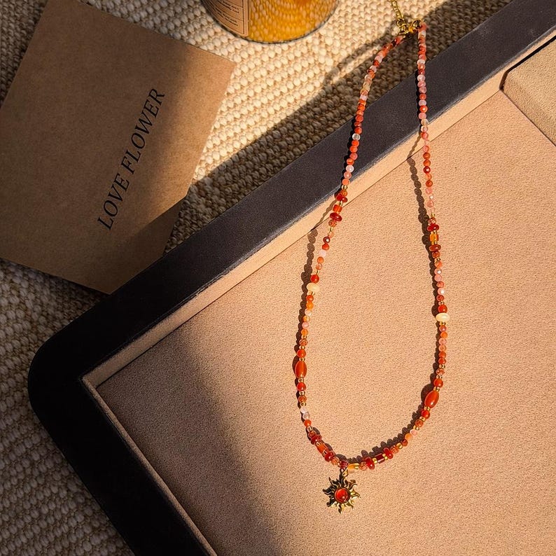 May include: A beaded necklace with a gold pendant in the shape of a sunburst. The necklace features a variety of orange, red, and cream-colored beads. The necklace is displayed on a beige surface, with a brown paper bag that reads "LOVE FLOWER".