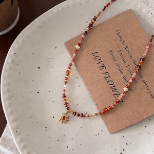May include: A beaded necklace with a sun pendant. The necklace features a mix of red, orange, and pink beads. The sun pendant is gold-colored with a red center. The necklace is displayed on a white plate with a brown card that reads "LOVE FLOWER".