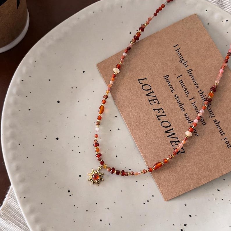 May include: A beaded necklace with a sun pendant. The necklace features a mix of red, orange, and pink beads. The sun pendant is gold-colored with a red center. The necklace is displayed on a white plate with a brown card that reads "LOVE FLOWER".