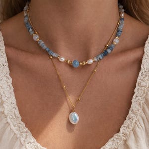 May include: A layered necklace featuring a gold chain with a large, white, teardrop-shaped pearl pendant. The upper layer has blue and white stone beads and small pearls. The necklace is worn against a white top.
