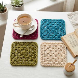 May include: Four square crochet coasters in magenta, teal, olive green, and cream. A white ceramic teacup with a silver spoon sits on the magenta coaster, while an open book and a small wooden lidded container are placed nearby on the table.