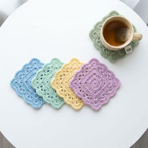 May include: A collection of handmade crochet coasters in pastel colors: blue, green, yellow, and purple. A cup of tea rests on a green coaster. The coasters have a square shape with a scalloped edge.