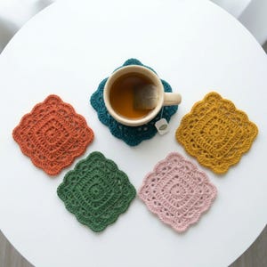 May include: Five square crochet coasters in orange, teal, dark green, mustard yellow, and light pink. A beige ceramic mug filled with tea and a tea bag sits on the teal coaster. Each coaster features a decorative scalloped edge and intricate stitching.