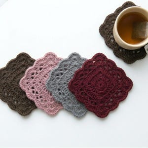 May include: A collection of handmade crochet coasters in various colors: brown, pink, gray, and burgundy. One coaster holds a beige mug with tea and a tea bag. The coasters are square with a scalloped edge.