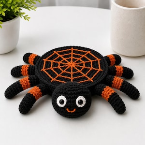 May include: A crocheted spider coaster featuring a round black body with an orange web pattern on top. The spider has eight legs with alternating black and orange stripes, a small round head with two white-rimmed black eyes, and a stitched orange smile.