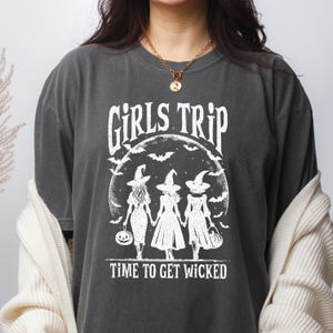May include: A charcoal gray short-sleeve t-shirt featuring a white graphic print of three figures in witch hats holding hands under a full moon with bats. The text reads GIRLS TRIP and TIME TO GET WICKED in a stylized white font on the front.