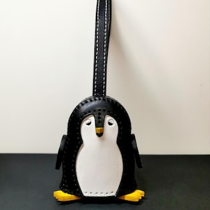 Può includere: Un portachiavi a forma di pinguino in pelle bianca e nera, con zampe gialle e becco giallo. Il portachiavi ha un cinturino nero con cuciture bianche. Il pinguino ha una pancia bianca e ali nere.