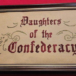 May include: Framed needlepoint with the words "Daughters of the Confederacy" in burgundy thread. The text is in a gothic font, with decorative green embroidered flourishes. The frame is dark brown.