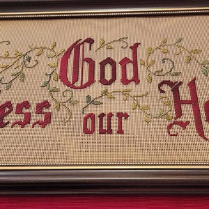 May include: A framed embroidery with the text "God Bless Our Home" in red lettering with green leafy vines surrounding the text.