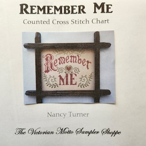 May include: Counted cross stitch chart pattern for a sampler with the text "Remember Me" in a vintage font with a small heart above the word "Me". The sampler is framed in a dark wood frame.
