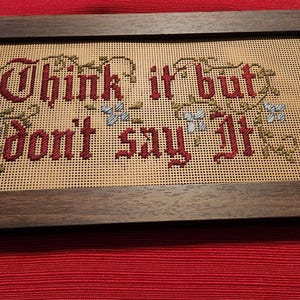 May include: A framed cross-stitch piece with the words "Think it but don't say it" in burgundy thread. The text is surrounded by floral accents in green and blue, set against a tan background. The frame is dark brown.