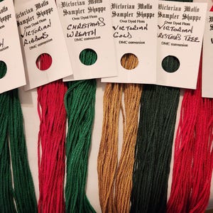 May include: A collection of over-dyed floss threads in various colors, including green, red, and gold, with attached labels. The labels read "Victorian Mollo Sampler Shoppe" and feature names like "Holly Branch" and "Christmas Wreath."