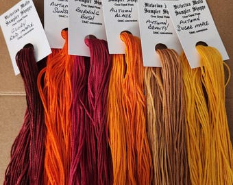 May include: A collection of colorful embroidery floss skeins in shades of red, orange, and yellow. Each skein is labeled with a name and DMC conversion. The colors range from deep burgundy to bright yellow, perfect for cross-stitch or embroidery projects.