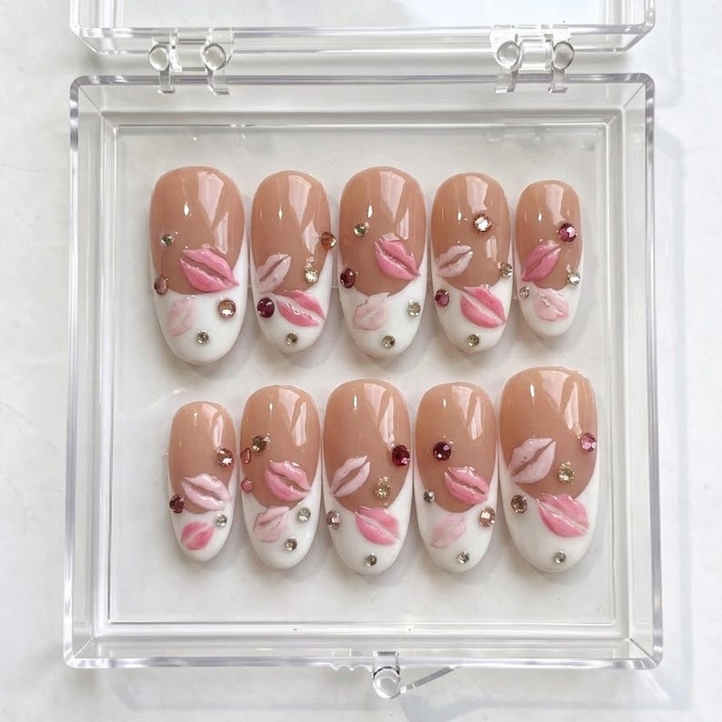 May include: A set of oval-shaped false nails in a clear plastic case. The nails feature a nude base with a white tip, adorned with pink lip designs and rhinestones. The nails are designed for a fashionable manicure.