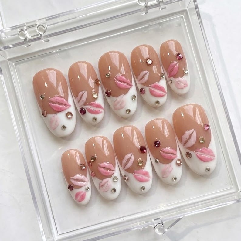 May include: A set of ten artificial nails in a clear plastic case. The nails are oval-shaped with a nude base and white tips. Each nail features a pink lip design, rhinestones, and a 3D effect.