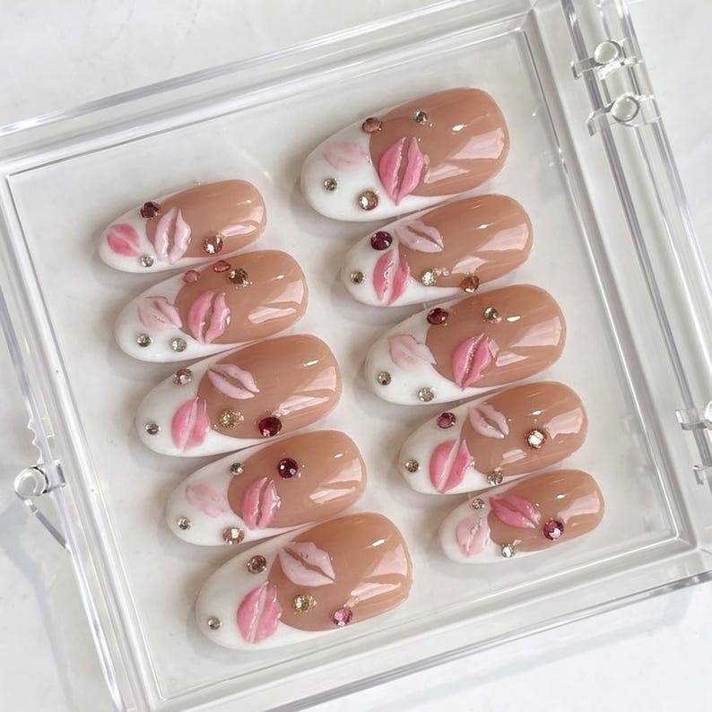 May include: A set of ten oval-shaped artificial nails. Each nail features a nude-colored base with a white tip, adorned with pink lip designs and sparkling rhinestones. The nails are arranged in a clear plastic case.