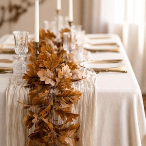 May include: A table runner featuring a garland of brown oak leaves draped down the centre of a white tablecloth. The table is set with white plates, clear textured glassware, and tall white taper candles in metallic holders for a dining display.