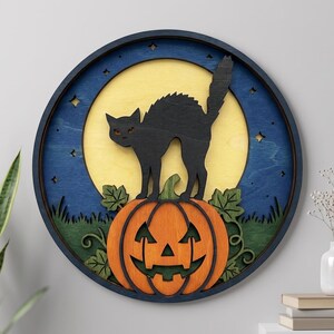 May include: A circular wooden wall art piece featuring a layered design of a black cat with an arched back standing on an orange jack-o'-lantern. The background displays a yellow full moon, a dark blue night sky with stars, and green foliage.