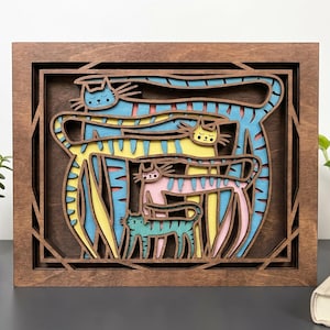 May include: A multi-layered wooden wall art piece featuring a group of stylized, elongated cats in shades of blue, yellow, and pink. The cats are arranged in a stacked composition within a dark brown wooden frame with geometric cutouts.
