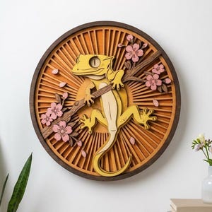 Op de afbeelding: Een cirkelvormig houten wandkunstwerk met een gele gekko die op een bruine tak met roze kersenbloesems klimt. De achtergrond bestaat uit een gelaagd zonnestraalpatroon in tinten oranje en bruin, omlijst door een donkere cirkelvormige rand.