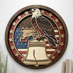 May include: A circular wooden wall art piece featuring a layered design of a bald eagle perched atop the Liberty Bell. The background displays a stylised American flag with red, white, and blue colours, surrounded by a border of carved wooden stars.