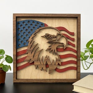 Op de afbeelding: Een vierkant houten wandkunstwerk met een gelaagde uitsparing van een Amerikaanse zeearendkop in donkerbruin hout. Achter de adelaar bevindt zich een gestileerd Amerikaans vlagontwerp met een blauw gedeelte met sterren en horizontale rode strepen.