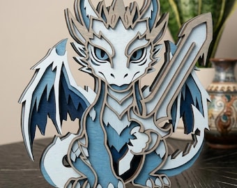 May include: A multi-layered wooden cutout of a dragon in shades of blue, white, and gray. The dragon features sharp horns, detailed wings, and holds a large sword in its claw. The design uses stacked wood pieces to create a three-dimensional effect.