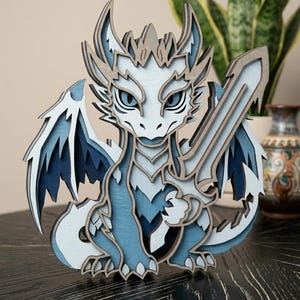 May include: A multi-layered wooden cutout of a dragon in shades of blue, white, and gray. The dragon features sharp horns, detailed wings, and holds a large sword in its claw. The design uses stacked wood pieces to create a three-dimensional effect.