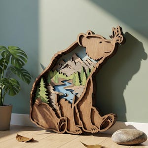 May include: A wooden bear-shaped wall art piece with a layered design. The bear has a butterfly on its nose and a landscape scene inside, including mountains, trees, and a river. The art is made of light brown wood.
