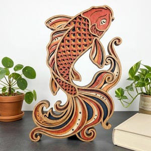 May include: A layered wooden koi fish sculpture in shades of orange, red, and brown. The fish is depicted swimming upwards with detailed scales and flowing fins. The sculpture is placed on a dark surface with plants and a book in the background.