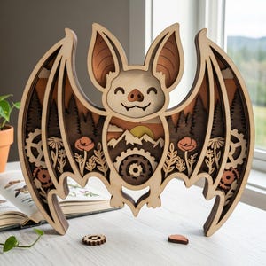 Peut inclure: Décoration murale en bois en forme de chauve-souris avec un design en couches. La chauve-souris présente un visage souriant, des ailes détaillées et une scène intérieure avec des montagnes, des arbres et des engrenages. La palette de couleurs comprend des tons de bois naturels, du marron et des accents orange.