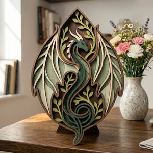 May include: A layered wooden dragon sculpture in shades of green, brown, and cream. The dragon is intricately cut with leafy accents and a wing design. The sculpture is displayed on a wooden table.
