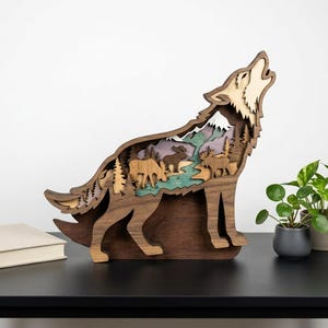 May include: A layered wooden sculpture of a howling wolf. The wolf's interior depicts a mountain scene with trees, animals, and a river. The sculpture is brown, with green, purple, and tan accents. A book and potted plants are nearby.