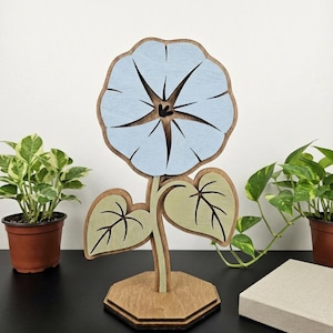 以下が含まれることがあります： 木製の花の彫刻。薄い青色の花と緑の葉があります。花は木の茎と台座に取り付けられています。花びらは星のようなデザインです。彫刻は装飾品です。