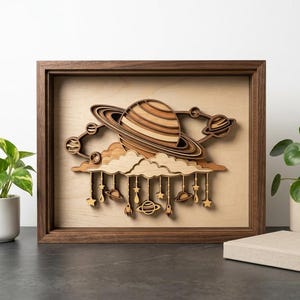May include: A framed wooden art piece featuring a layered design of a planet with rings, planets, clouds, stars, and rockets. The artwork is in a dark brown frame and is displayed on a dark surface with plants and a book.