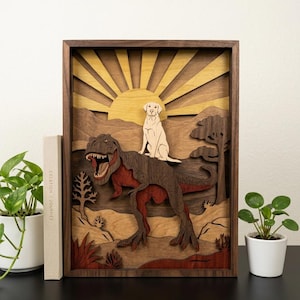 Op de afbeelding: Houten kunstwerk met meerdere lagen, met een Tyrannosaurus Rex met een hond op zijn rug, tegen een zonsondergang. Het kunstwerk is ingelijst in donker hout, met een gedetailleerd, meerlagig ontwerp. De hond is crème van kleur.