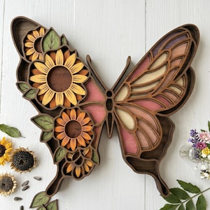Peut inclure: Décoration murale en bois en forme de papillon avec un design en couches. L'aile gauche présente des tournesols dans des tons de jaune et d'orange, avec des feuilles vertes. L'aile droite a des tons roses, pêche et bruns. Le papillon est posé sur un fond blanc.