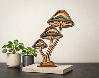 Puede incluir: Una escultura de hongos de madera en capas con tres hongos de diferentes tamaños. Los sombreros de los hongos presentan capas de colores naranja, verde y crema. La escultura está sobre un libro con el texto "LAYERED ARTISTRY".