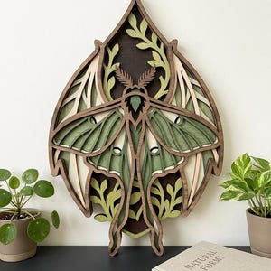 May include: A multi-layered wooden wall art piece shaped like a pointed leaf, featuring a central moth design in shades of dark brown, sage green, and cream. The intricate laser-cut layers create depth, showcasing detailed wings and foliage patterns.