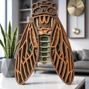 May include: A layered wooden cicada sculpture with intricate cut-out details. The insect features a dark brown head, tan wings, and a green and tan body. The artwork is displayed on a white table, showcasing a modern home decor piece.