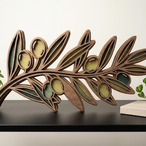Puede incluir: Escultura de rama de olivo de madera con diseño en capas. Las hojas y aceitunas están pintadas en tonos verdes y amarillos, sobre un fondo de vetas de madera marrón. La pieza decorativa está colocada sobre una superficie negra.