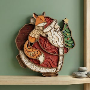 May include: A layered wooden Christmas decoration featuring Santa Claus carrying a Christmas tree and a fox wearing a sweater. The Santa figure is red and white, with a brown bag. The fox is orange and white.