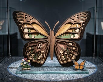 May include: A large, intricately carved wooden butterfly sculpture with layered wings, showcasing a forest scene. The wings are a mix of brown, tan, and green, with a small butterfly and a floral arrangement at the base. A smaller butterfly model is also present.
