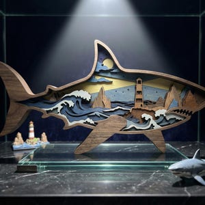 May include: A wooden shark sculpture with a layered coastal scene design. Inside the shark, there's a lighthouse, waves, and a moonlit sky. The shark is displayed on a glass surface, alongside a small shark figurine and a lighthouse model.