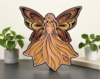 May include: A layered wooden fairy sculpture with a detailed design. The fairy has large wings and a flowing dress, with colors ranging from dark brown to light peach and yellow. The artwork is displayed on a dark surface.