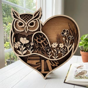 Pode incluir: Arte de parede em madeira em forma de coruja com uma moldura em forma de coração. O design em camadas apresenta uma coruja, flores e livros. A obra de arte é feita de madeira castanha clara e escura, criando uma estética natural e rústica. A peça é exibida em uma superfície branca.